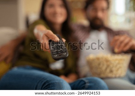 Close-up of a remote control held by a woman with a couple relaxing on a couch, eating popcorn and watching TV at home.