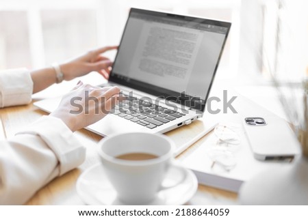Close-up of the right female hand. Unrecognizable woman is typing text on a laptop. Cup of coffee, smartphone, glasses on the desktop. Blurred foreground.