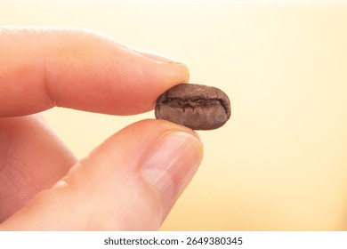 Close-up of a roasted coffee bean, split open, held by a hand against a light beige background. A perfect shot for coffee culture, barista, or beverage-related projects.