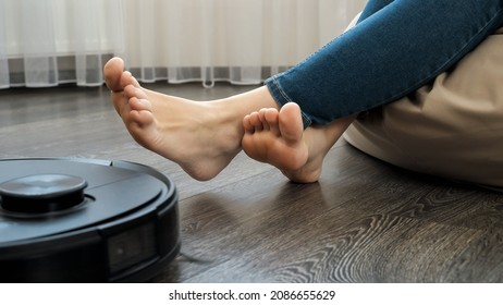 Closeup Of Robot Vacuum Cleaner Driving Around Barefoot Woman Working At Home Office. Concept Of Hygiene, Household Gadgets And Robots At Modern Life.