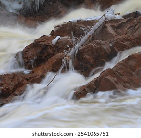 A close-up of a rocky waterfall. Water rushes over dark, wet rocks, creating a white, frothy cascade. A fallen tree branch lies across the top of the falls, adding a touch of contrast to the scene. - Powered by Shutterstock