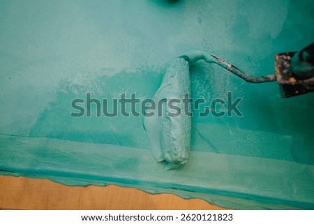 Close-up of a roller spreading thick blue waterproofing sealant on a bathroom floor, with visible texture and edge of the plywood surface.