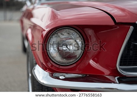 Close-up of a round headlight on a red classic American muscle car with a shallow depth of field, highlighting iconic automotive design and vintage chrome details.