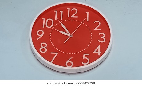 Close-up of a round red wall clock with a white frame and white numerals mounted on a light blue wall. The clock displays approximately 09:54 and 2 seconds, with clear hour, minute, and second hands. Concept of time management, schedule, punctuality, daily routine, and office or home interior.
