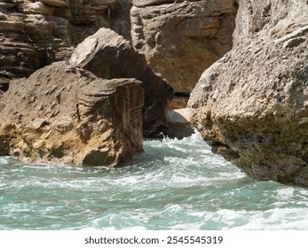 Close-up of rugged coastal rocks with ocean waves crashing against them. The rough textures of the stone contrast with the movement of the turquoise water below. - Powered by Shutterstock