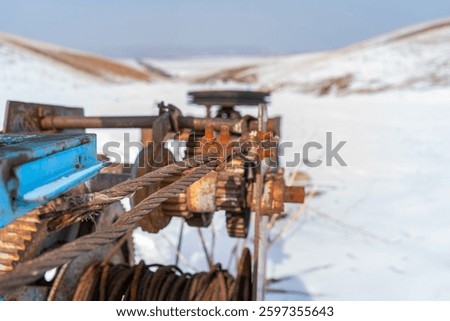A close-up of a rusted cable winch set against a snowy hillside, revealing weathered metal gears and bolts. The stark winter setting highlights the machinery’s rugged character