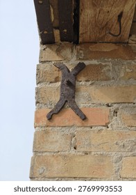 Close-up of a rustic brick wall, aged and weathered, featuring a prominent, heavily rusted metal X-shaped bracket secured by a bolt. An old wooden beam with a bent hook is nearby, against a bright