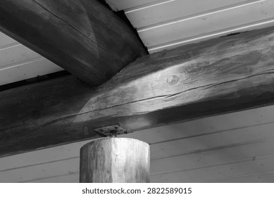 Close-up of a rustic log beam ceiling and round support post with visible timber grain, knots and metal bracket. Monochrome interior detail emphasizing texture, craftsmanship, structural stability 