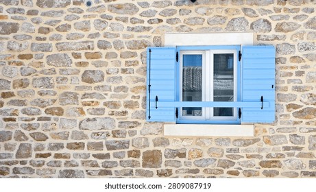 Close-up of a rustic stone wall with a white-framed window featuring light blue wooden shutters and a crossbar, showcasing traditional Mediterranean architecture.