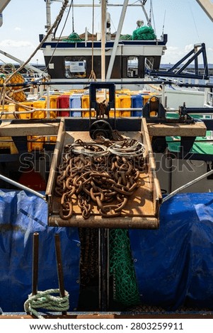 Close-up of rusty iron anchor chains and steel cables piled in a metal bin mounted on the deck of a fishing trawler. Colorful fish transport crates visible in the background on deck.