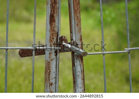 Close-up of a rusty iron gate secured with a padlock. Symbol of security, abandonment, or restricted access. Rural setting with blurred green background.
