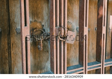 Close-up of rusty padlock and chain securing old wooden door with metal bars. Weathered wood texture, corroded iron details and vintage hardware. Authentic aged security element with rich brown and orange rust tones.