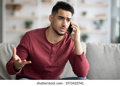 Closeup Of Sad Arab Guy Sitting On Couch, Having Conversation On Mobile Phone. Angry Middle-eastern Young Man Talking On Smartphone, Fighting With Girlfriend Or Customer Service