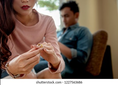 Close-up of a Sad Wife want to Divorce Lamenting Holding the Wedding Ring in a House Interior with Blurred Man on Sofa Background.