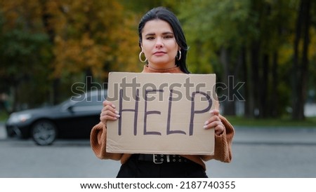 Close-up sad young woman stands outdoors near roadway holding cardboard sign with words HELP upset hispanic girl searching for job experiencing financial crisis problems of dismissal unemployment