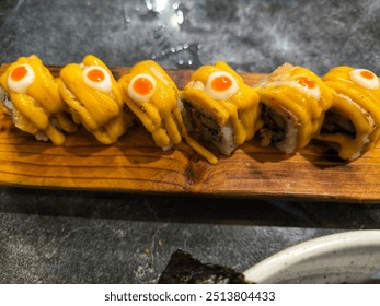 Close-up of a salmon sushi roll drizzled with a creamy yellow sauce and topped with a spicy red sauce, served on a wooden board. - Powered by Shutterstock