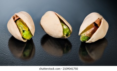 Closeup of salty roasted pistachio nuts cores with reflection on shiny black background. Pistacia vera. Three beautiful green pistachios kernels in beige split halved shells on rough textured surface.