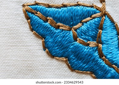 Close-up of satin stitch embroidery of floral ornament on a cotton white tablecloth. A bright blue embroidered flower is framed with beige threads. Needlework, DIY concept. Top view, macro, flat lay