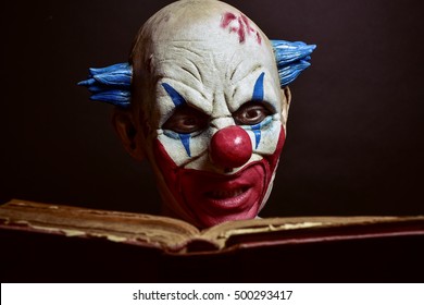 Closeup Of A Scary Evil Clown Reading An Old Book, Against A Dark Background