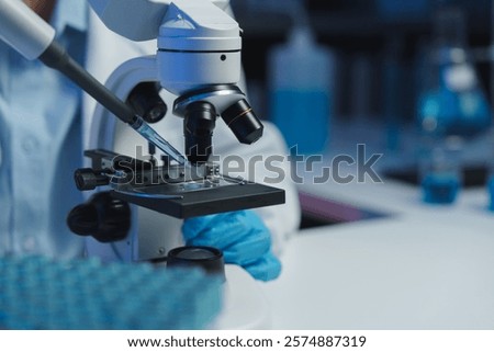 Close-up of a scientist in gloves placing a sample on a microscope slide with a pipette, preparing for analysis in a modern lab filled with blue liquids