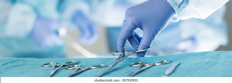 Close-up of scrub nurse taking medical instruments for operation with colleagues performing in background. Letter box format