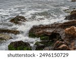 Close-up of sea waves hitting rocky shore, showcasing foam, surf, and moss-covered stones in marine environment.