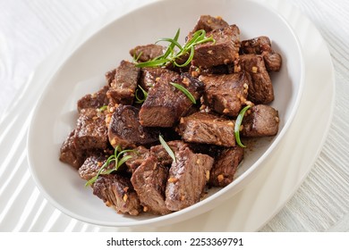 close-up of seared juicy new york strip steak bites with butter, rosemary and garlic in bowl on white wood table