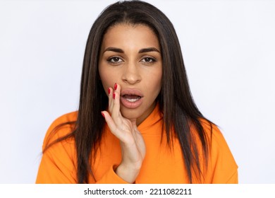 Closeup Of Secretive Hispanic Woman Whispering. Confident Young Model With Long Hair In Orange Hoodie Looking At Camera, Speaking With Hand Near Mouth. Secret, Rumor Concept.