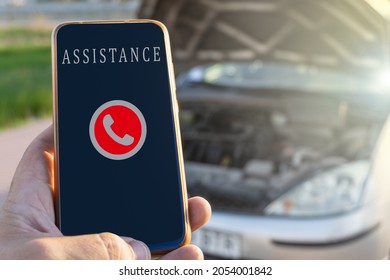 Close-up with selective focus of a phone ready to call the roadside assistance service, with a damaged car in the background.