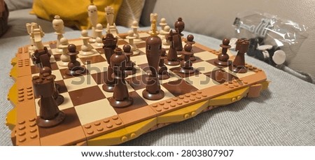 Close-up selective focus shot of a chess board and chess pieces entirely constructed from plastic building bricks. The brown and beige pieces, including the king, queen, rooks, and pawns, are arranged on a checkered brick board over a soft indoor couch background. This image perfectly illustrates strategy, logic, intellect, board games, building block hobbies, smart entertainment, and educational activities for kids and adults.
