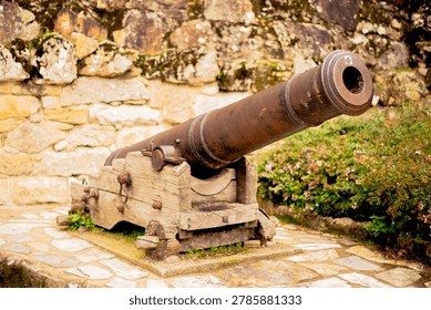 A close-up, selective focus shot of a rusty vintage cannon mounted on a traditional wooden carriage. Set against a stone wall background with flowers, this historical artillery piece represents military heritage and ancient fortifications.