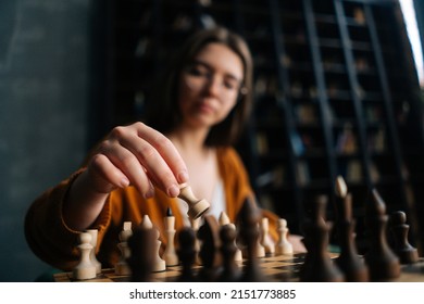 Close-up selective focus of smart young woman in elegant eyeglasses making chess move sitting in armchair in dark library room. Pretty intelligent lady playing logical board game alone at home.