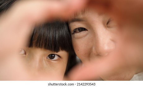 Closeup With Selective Focus Smiling Asian Mom And Daughter Making Heart Gesture With Hands While Taking Selfie. Love And Family Concept