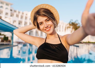 Close-up selfie-portrait of attractive girl with long hair standing near pool. She is smiling to the camera and shows cool look. Straw hat on head.  On resort, holiday, vacation.