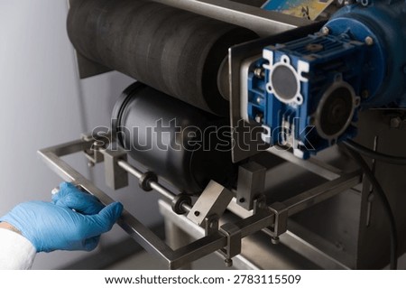 Close-up of a semi automatic labeling machine processing a black plastic supplement container in a modern nutraceutical manufacturing facility. Industrial worker operating packaging equipment for dietary supplements and whey protein products. Concept of food industry automation, private label production, pharmaceutical packaging, quality control, and industrial manufacturing technology.