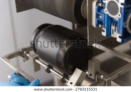 Close-up of a semi automatic labeling machine processing a black plastic supplement container in a modern nutraceutical manufacturing facility. Industrial worker operating packaging equipment for dietary supplements and whey protein products. Concept of food industry automation, private label production, pharmaceutical packaging, quality control, and industrial manufacturing technology.