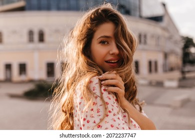 Close-up of semi-sideways beauty caucasian young girl posing in blouse on blurred background of city. Light blond hair covers half of her face. Stylish woman having good time.