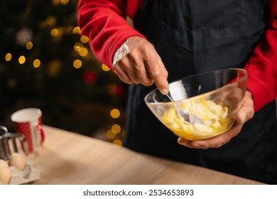 Close-up of senior man mixing ingredients for Christmas cookie dough in a glass bowl. The man is wearing festive clothing and working in a Christmas-themed kitchen. - Powered by Shutterstock