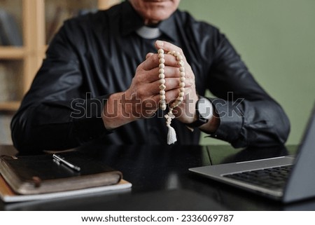 Closeup of senior priest holding rosary while praying at desk in office, copy space