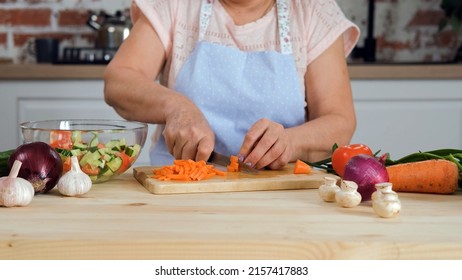 Closeup Of Senior Woman In Apron Chopping Carrots On The Wooden Board In The Kitchen. Slow Motion. Cooking And Healthy Vegan Eating Concept