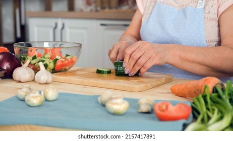 Closeup Of Senior Woman In Apron Chopping Cucumbers On The Wooden Board In The Kitchen. Slow Motion. Cooking And Healthy Vegan Eating Concept