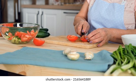 Closeup Of Senior Woman In Apron Cutting Tomatoes On The Wooden Board In The Kitchen. Slow Motion. Cooking And Healthy Vegan Eating Concept