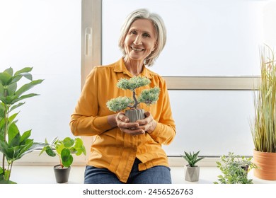 Closeup, senior woman holding bonsai tree, caring to home flower pot at balcony. Attractive pensioner growing plant, handmade. Home hobby concept - Powered by Shutterstock