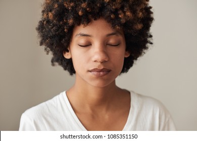 Closeup of a serene young African woman with an afro and natural complexion standing with her eyes closed against a gray background