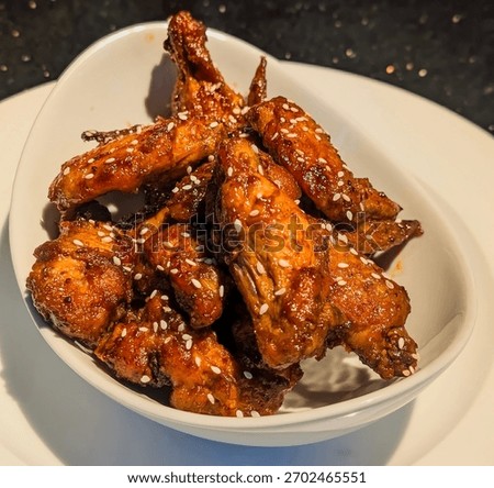 A close-up of a serving of deeply glazed, sticky BBQ or spicy chicken wings, generously coated in a rich brown sauce and sprinkled with white sesame seeds.