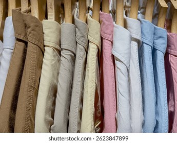a close-up of several collared shirts in an array of soft, light, and earthy colors. The shirts, which exhibit a visible fabric texture, are neatly arranged on wooden hangers. 