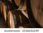 A close-up of several wooden wine barrels stacked in a cellar. The barrels are made of dark brown wood with silver metal hoops. The image has a warm, rustic feel.