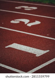 Close-up sharp focus shot of the white starting line and '1' lane marker on a vibrant red synthetic athletic running track surface.