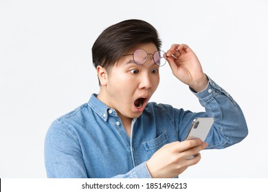Close-up of shocked asian man take-off glasses, gasping in awe while looking at mobile phone screen, reading shocking news, react to announcement on smartphone, white background