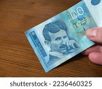 A close-up shot of a 100 Serbian dinar banknote with the image of the famous scientist Nikola Tesla. A person holds a banknote in his hand